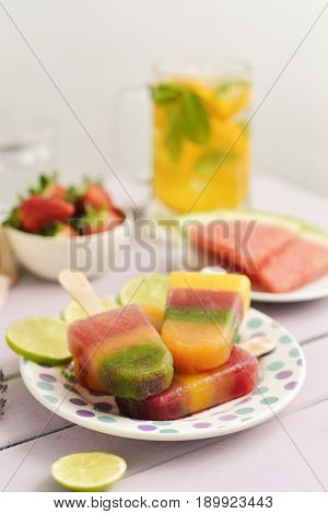 Homemade natural ice pops in a ceramic plate next to pieces of fruit, such as strawberries or watermelon, on a rustic pink table