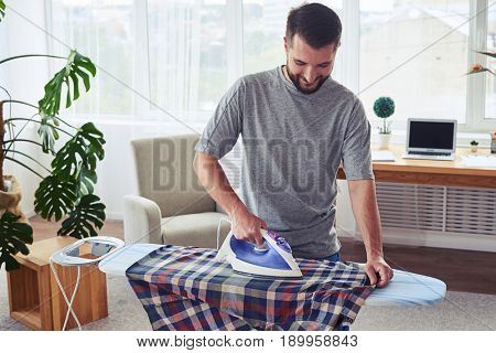 Mid shot of man with charming smile ironing diligently shirt on ironing board