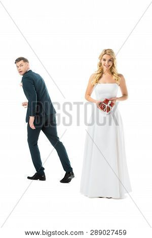 Smiling Pretty Bride Holding Gift Box, While Tricky Groom Imitating Running Away Isolated On White