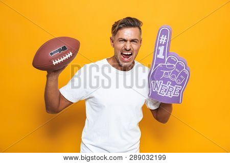 Image of cheerful man 30s in white t-shirt holding rugby ball and number one fan hand glove with finger raised while standing isolated over yellow background