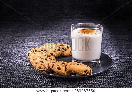 A Glass Of Milk With Cinnamon And Chocolate Chip Cookies. On A Black Plate On A Gray Tablecloth.