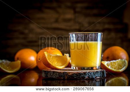 A Glass Of Fresh Orange Juice On A Wooden Board And A Table With Orange Slices On A Stone Wall Backg