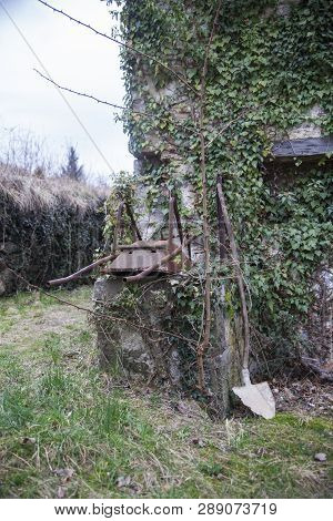 Old And Rusty Wheelbarrow And Shovel On A Wall Of A House Overgrown With Poison Ivy In Abandoned Vil