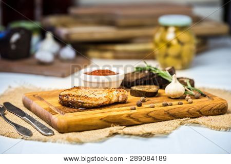 Pork Chop Lies On A Board With Spices, Garlic, Rye Bread, Baked Vegetables And Sauce. Blurred Backgr