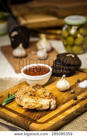 Pork Chop Lies On A Board With Spices, Garlic, Rye Bread, Baked Vegetables And Sauce And Blurred Bac
