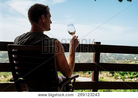 A Handsome Adult Man Sits On A Farm And Tastes A Glass Of White Wine. Sunny Day, Cyprus.