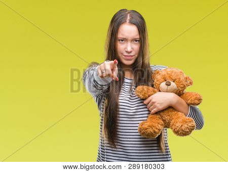 Young caucasian woman holding teddy bear over isolated background pointing with finger to the camera and to you, hand sign, positive and confident gesture from the front