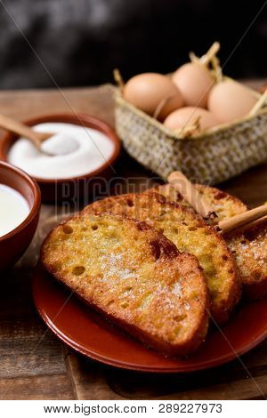 an earthenware plate with some torrijas, typical spanish dessert eaten in Lent and Easter, and some of the ingredients to prepare it, such as eggs, cinnamon or sugar, on a wooden table