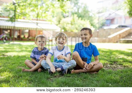 Three Happy Young Boys In Summer Park. Friends Or Siblings Sitting On Green Grass.