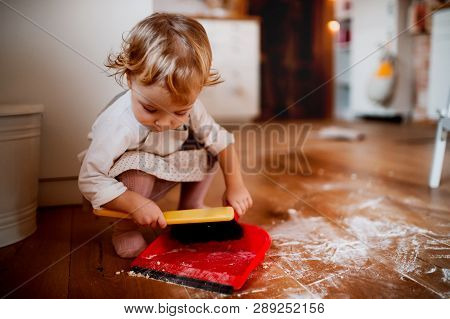 A Small Toddler Girl With Brush And Dustpan Sweeping Floor In The Kitchen At Home.