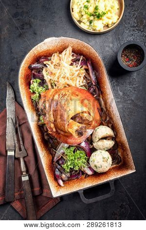 Traditional Bavarian schweinshaxe with onion, potato salad and sauerkraut as top view in a rustic stewpot on an old board
