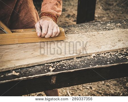 Carpenter In Medieval Cotton Clothes Working With A Wood By Plane. Man Manually Hews A Wooden Board 