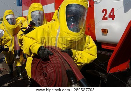 Firefighters In Protective Suits, Gas Masks And A Hose Are Getting Ready For Work. Mechanisms And De
