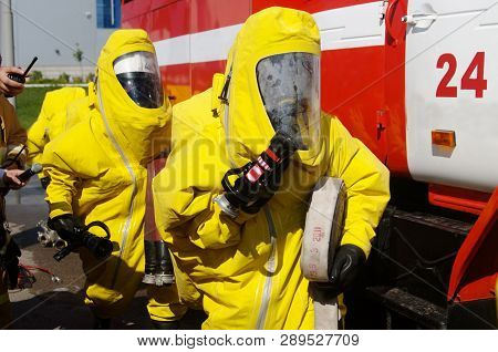 Two Firefighters In Protective Suits And Gas Masks Are Preparing For Work. Mechanisms And Devices Fo
