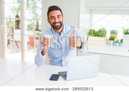 Handsome hispanic man working using computer laptop and drinking a cup of coffee happy with big smile doing ok sign, thumb up with fingers, excellent sign