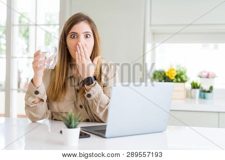 Beautiful young woman working with computer takes a break to drink glass of water cover mouth with hand shocked with shame for mistake, expression of fear, scared in silence, secret concept