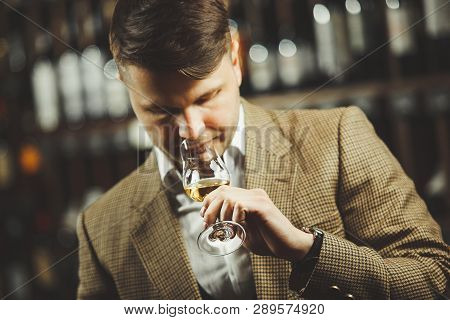 Sommelier Smelling Flavor Of Whiskey In Bokal On Background Of Shelves With Bottles In Cellar.