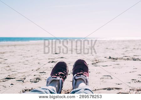 Feet Of Young Woman In Sport Footwear And Jeans Relaxing After Hiking On Sand Beach. Pov. Adventure 