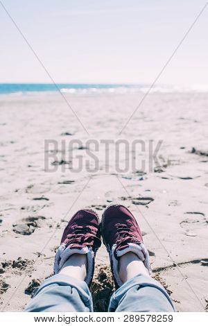 Feet Of Young Woman In Sport Footwear And Jeans Relaxing After Hiking On Sand Beach. Pov. Adventure 