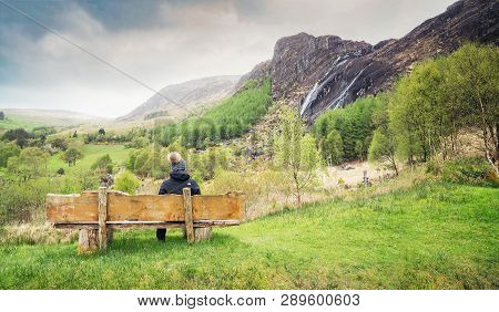 A Girl Sitting On The Wooden Bench And Looking At The Waterfall In Kenmare