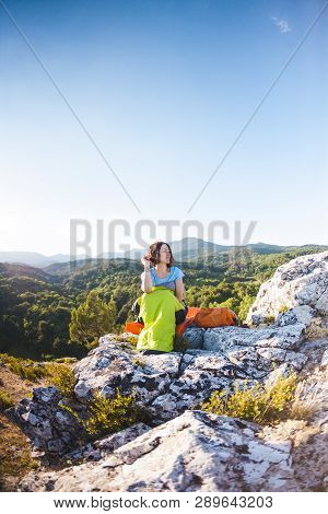 A Woman Is Sitting In A Sleeping Bag At Sunset. Camping On Top Of A Mountain. A Smiling Girl Rests A