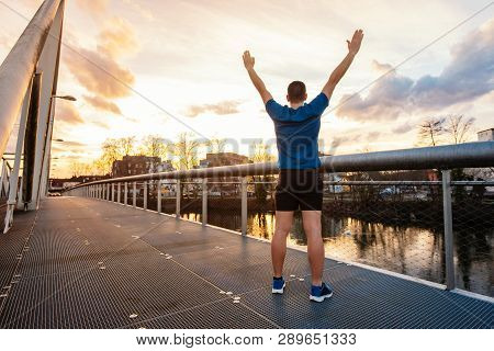 Rear View Of Young Man Winner Celebrating Sport Success As Raising Hands Up Standing On A Bridge Ove