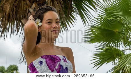 Young Girl With Floral Sundress Smiling With Flower In Hair Standing Under Palm Tree