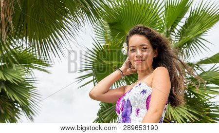 Young Girl With Floral Sundress Smiling With Flower In Hair Standing Under Palm Tree