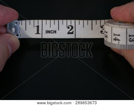 Macro Extreme Close-up Of Man Fingers Holding Measure Tape Ruler With Few Inches Unrolled On Dark Bl