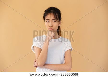 Portrait of obused asian woman wearing basic t-shirt posing at camera with angry look isolated over beige background in studio