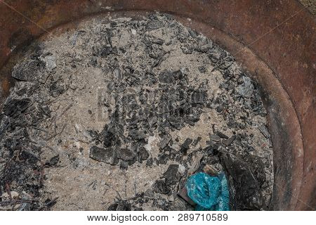 Closeup View Inside Metal Fire Pit Containing Ash, Charred Wood, Loose Nails. And An Unburnt Blue Pl