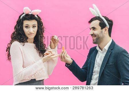 Young Happy Couple On The Pink Background. On The Head Is A Rabbit Ears. A Young Man Holding A Carro
