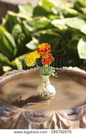 Small Vase Holding Marigolds Sitting In The Center Of A Birdbath On A Hot, Sunny, Summer Day.