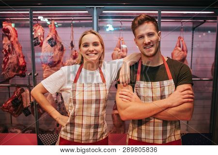 Young Man And Woman Stand At Meat Shelf In Grocery Store. They Pose On Camera And Smile. Animal Braw