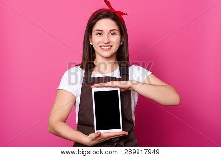 Photo Of Young Attractive Female Stands With Toothy Smile And Shows New Gadget Isolated Over Pink Ba