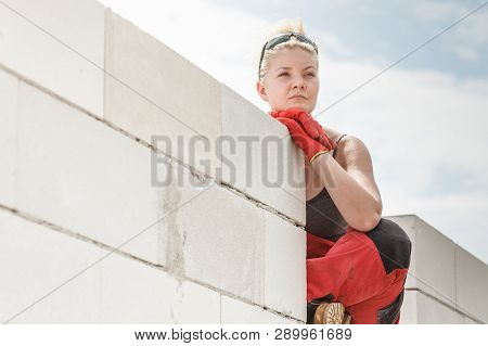 Woman Wearing Dungarees Thinking About Future Of Building Her Home. Female Sitting On Wall Made Of A