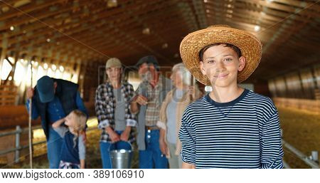Portrait Of Caucasian Small Teen Boy In Hat Standing In Shed And Smiling To Camera. Family On Backgr
