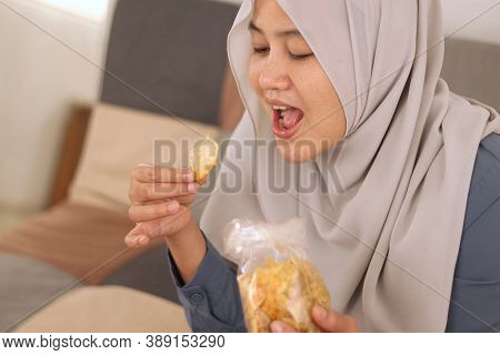 Asian Muslim Woman Enjoys Eating Casava Or Potato Chips While Sitting On Sofa At Living Room, Close 