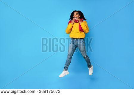 Excited Black Woman In Winter Jacket Jumping In Mid-air Looking Aside Posing Over Blue Studio Backgr