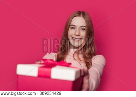Charming Ginger Woman With Freckles Is Giving A Present At The Camera Smiling On A Pink Wall At Stud