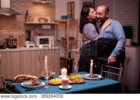 Kissing Husband Cheek While Greeting Him In Kitchen For Romantic Dinner. Woman Preparing Festive Din