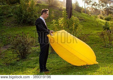 A Young Man Inflates A Yellow Lamzac With Air. A Man Inflates An Air Sofa In Park.