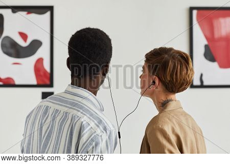 Back View Portrait Of Two Young People Looking At Paintings And Sharing Audio Guide While Exploring 