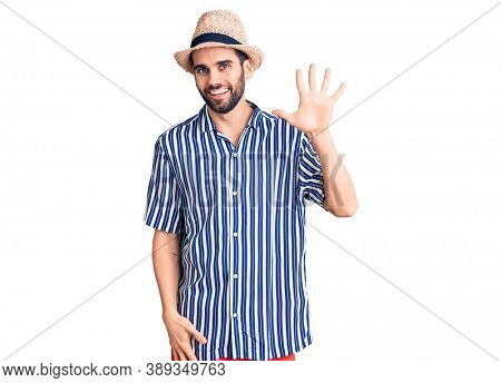 Young handsome man with beard wearing summer hat and striped shirt showing and pointing up with fingers number five while smiling confident and happy. 