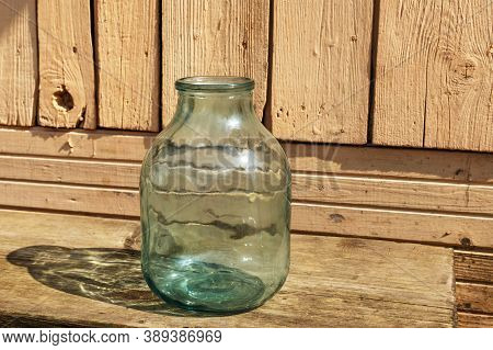Empty Glass Jar. There Is A Three-liter Jar On A Wooden Bench Near The Wall Of The House.