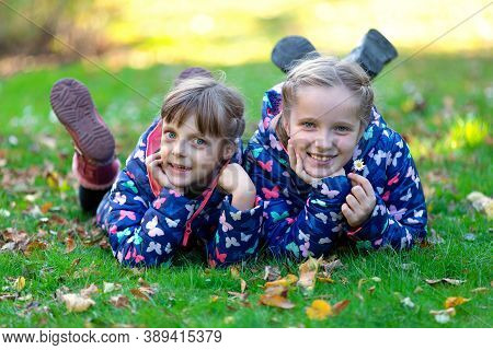 Two Smiling Kids Siblings Girls Lying On Green Grass With Autumn Fallen Leaves