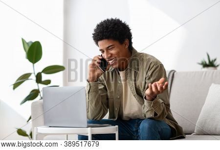 Elearning Concept. Positive Black Teenager Speaking On Cellphone Near Laptop Computer At Home. Afric