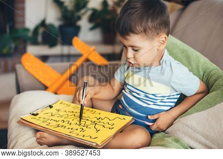 Lovely Brunette Boy Drawing On A Yellow Paper Using A Marker While Sitting On Pillows At Home During