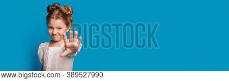 Small Caucasian Girl Is Gesturing The Stop Sign And Smile At Camera While Posing On A Blue Studio Wa