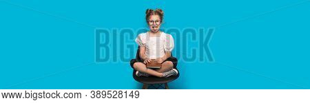 Caucasian Girl With Glasses Is Smiling And Showing Tongue While Sitting In Armchair Holding A Book O